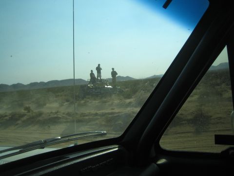 1990 Ford Bronco - Three silhouetted spectators stand atop a parked vehicle in the desert, viewed through a chase truck's windshield.