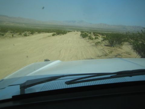 1990 Ford Bronco - faint two-track desert road stretches toward distant mountains, seen over the white hood of a moving vehicle.