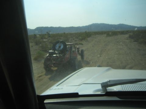 1990 Ford Bronco - red open-frame off-road buggy sits off the trail in the desert, viewed through a dusty chase vehicle window.