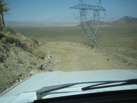1990 Ford Bronco - rocky desert two-track trail descends toward a large power-line tower, seen over the hood of a moving vehicle.