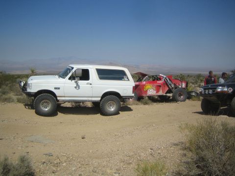 1990 Ford Bronco - white full-size Ford Bronco with a brush guard sits beside a disabled red race buggy on a desert trail.