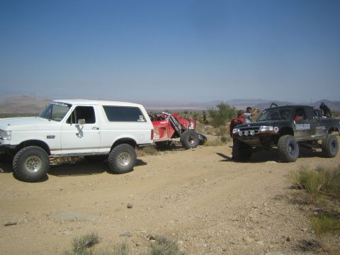 1990 Ford Bronco - white Ford Bronco and a dark prerunner truck flank a stranded red race buggy on a desert dirt road.