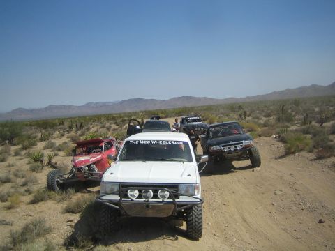 1990 Ford Bronco - white off-road truck marked The Web Wheeler leads a red buggy and dark prerunner along a desert two-track.