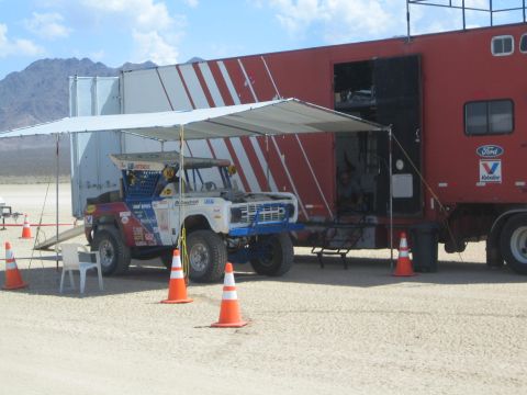 1990 Ford Bronco - race buggy sits under an awning beside a red Ford and Valvoline branded support truck in a desert pit area.