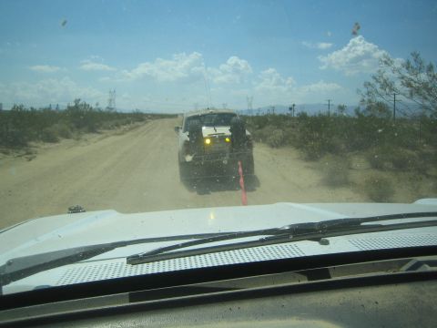 1990 Ford Bronco - dark off-road vehicle with amber lights drives ahead on a dusty desert road, viewed over a white vehicle hood.