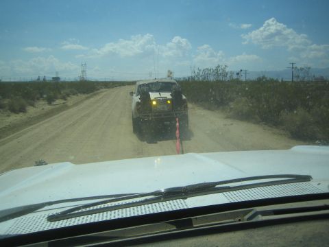 1990 Ford Bronco - muddy off-road vehicle with amber turn lights heads down a wide desert dirt road under a partly cloudy sky.