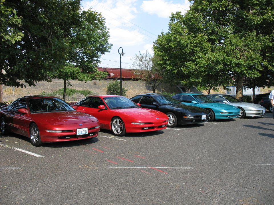Five Ford Probe cars parked in a lot, with trees and a train in the background
