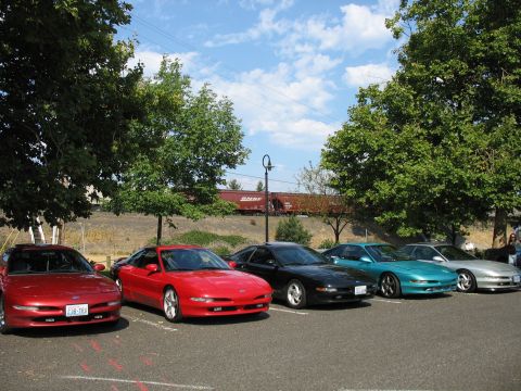 1997 Ford Probe - A line of Ford Probes are parked in a lot under trees, with a train passing by in the distance.