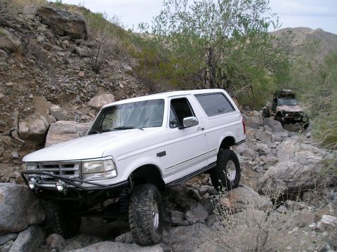 1993 Ford Bronco - A white 1993 Ford Bronco tackles a challenging rocky trail, with another off-road vehicle following behind.