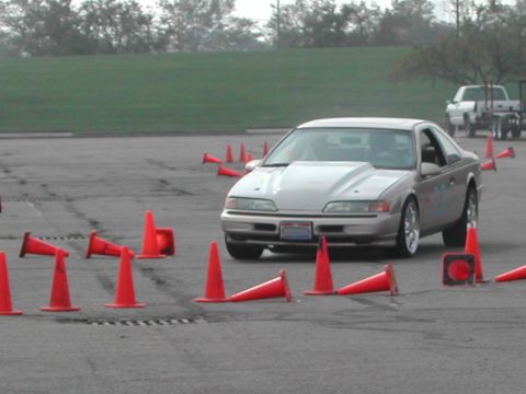 1993 Ford Thunderbird - A silver 1993 Ford Thunderbird navigates an autocross course marked by orange cones, with a white pickup truck visible in the background.