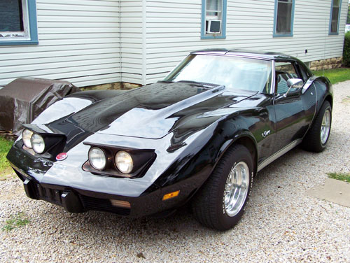 Black C3 Corvette coupe, front three-quarter view with headlights up, on gravel driveway