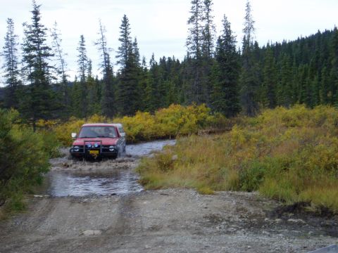 2000 Ford E350 - Red SUV navigates a water crossing in a wooded area.