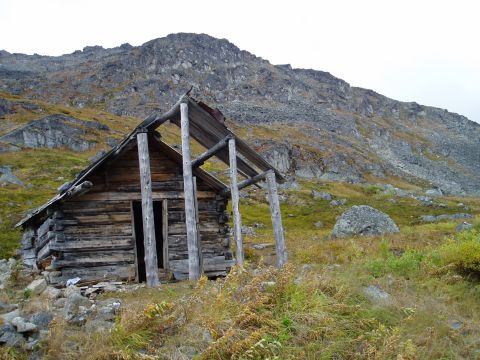 2000 Ford E350 - An old, weathered log cabin sits in a grassy, mountainous area, with its roof in disrepair.