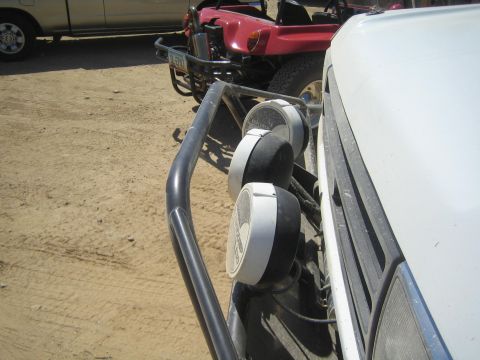 1990 Ford Bronco - Close-up of three round driving lights mounted on a white truck's tube brush guard with a red buggy parked behind.