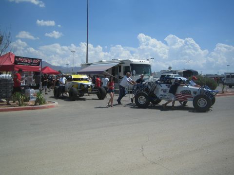 1990 Ford Bronco - Two tube-frame off-road race buggies, one yellow and one blue with a flag wrap, sit in a paved pit lot beside vendor tents and an RV.