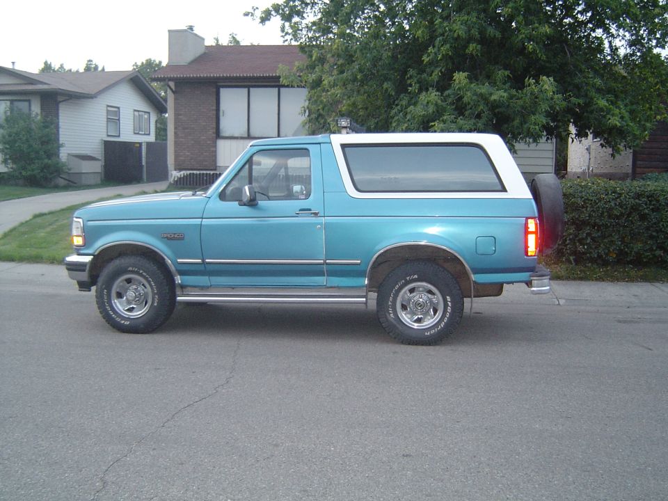 Blue and white Ford Bronco parked on a residential street. Side view shows chrome running boards and a spare tire mounted on the rear