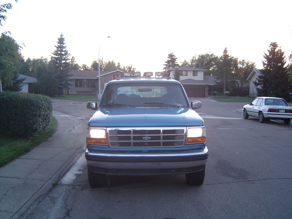 Blue Ford Bronco parked on street, front view. White sedan in background