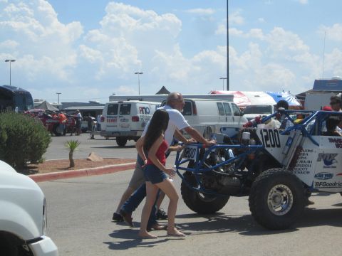 1990 Ford Bronco - man and woman push a blue number-200 tube-frame off-road race car across a paved pit area near vans and tents.