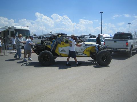 1990 Ford Bronco - People push a yellow and white number-108 tube-frame off-road race car past an RV and a white pickup in the pit area.