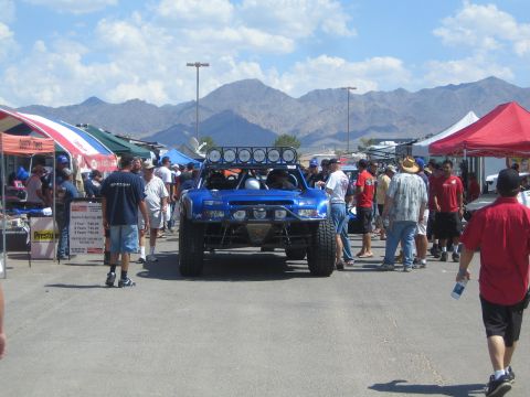 1990 Ford Bronco - blue off-road trophy truck is parked among a crowd of spectators and vendor tents with rugged mountains in the background.