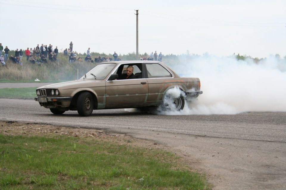 Tan two-door coupe doing a burnout with heavy white tire smoke as spectators watch