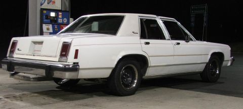 1985 Ford LTD Crown Victoria - A white sedan is parked at a gas station at night, viewed from the rear.