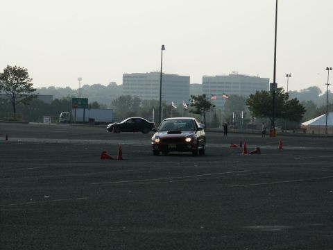 2001 Mercury Grand Marquis - A red Subaru WRX navigates a cone course in a large parking lot with buildings in the distance.