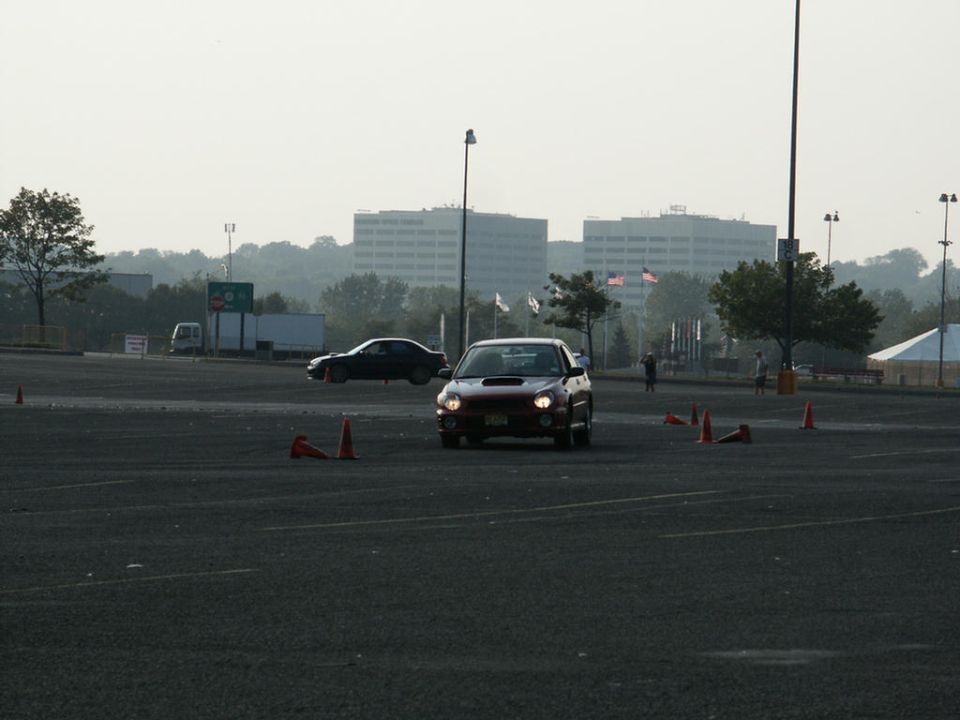 Red Subaru Impreza WRX driving through cones in a parking lot, another car in background