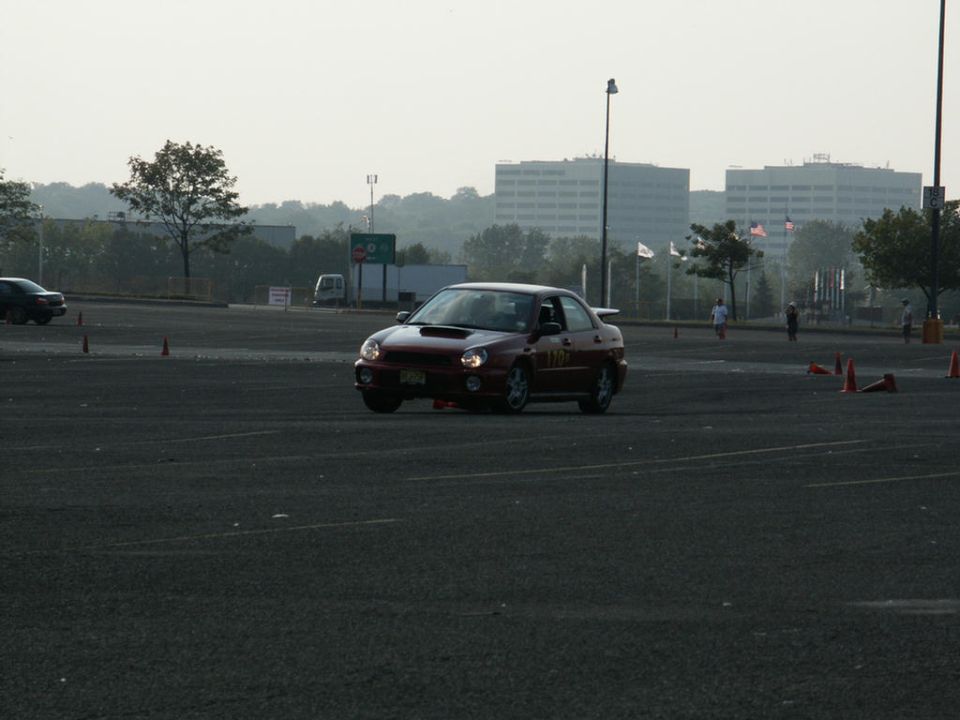 Red sedan with a spoiler and racing numbers on the door, angled from the front-right, driving on a dark asphalt lot with orange cones