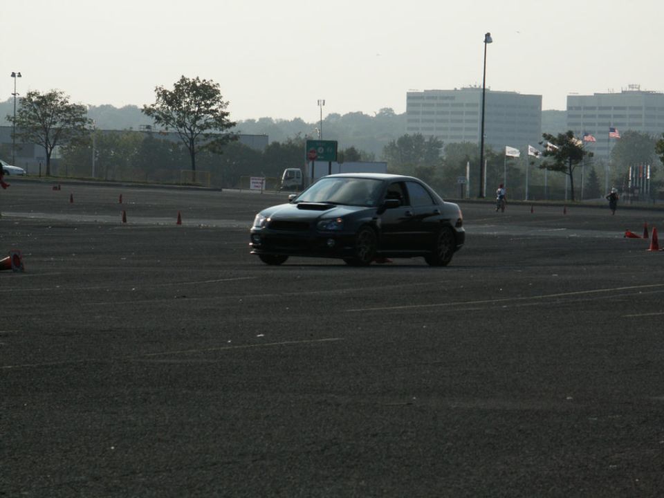 Black sedan with a hood scoop driving on a paved lot with orange cones