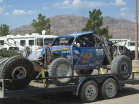 1990 Ford Bronco - blue number-519 tube-frame off-road race car sits chained on a tandem-axle trailer with RVs and desert hills behind.