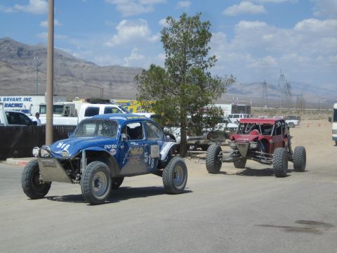 1990 Ford Bronco - blue number-518 Baja-style Volkswagen Beetle and a red tube-frame buggy sit on a dirt staging area with power lines and mountains behind.