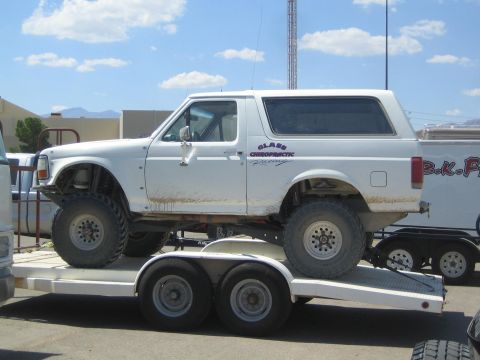 1990 Ford Bronco - white two-door full-size SUV with off-road tires and Glass Chiropractic Racing lettering sits on a tandem-axle flatbed trailer.