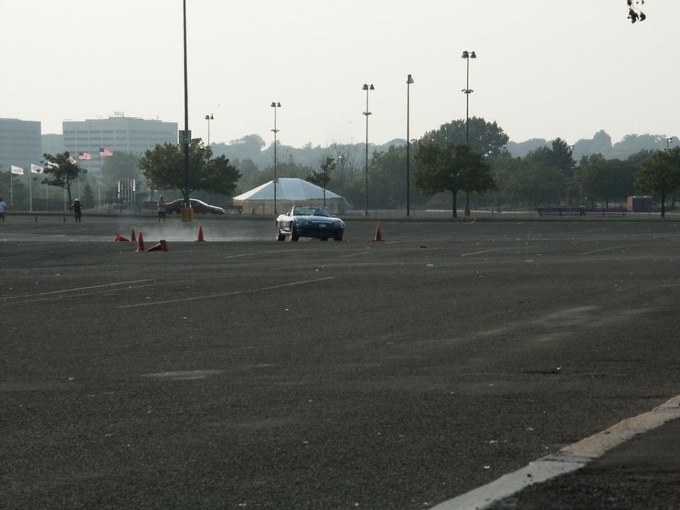 Blue convertible sports car on asphalt with orange cones, distant buildings and trees under hazy sky