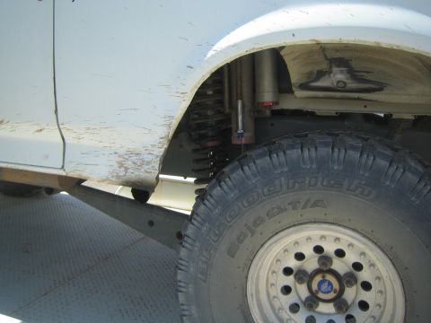 1990 Ford Bronco - Detail shot of a white off-road vehicle's front wheel well showing a coilover shock and a muddy BFGoodrich tire on a polished wheel.