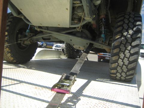 1990 Ford Bronco - Low-angle view under an off-road vehicle on a flatbed, showing the chassis, blue shocks and a ratchet tie-down strap.