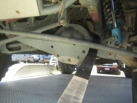 1990 Ford Bronco - Under-vehicle view showing an off-road truck's front axle, blue coilover shock and a black tie-down strap on a trailer deck.