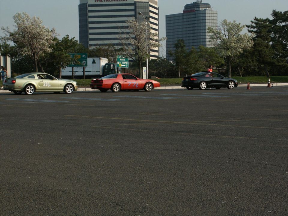 gold Mustang, red Camaro, and black coupe parked in a lot with buildings in the background