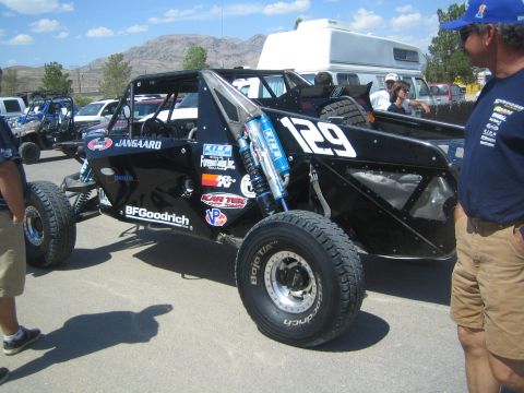 1990 Ford Bronco - black number-129 tube-frame single-seat off-road race buggy sits in a desert pit area with spectators and an RV nearby.