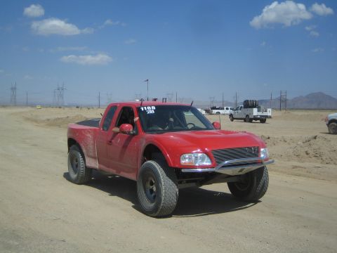 1990 Ford Bronco - red off-road prerunner pickup with fender flares drives on a dirt course with power lines and desert mountains behind.