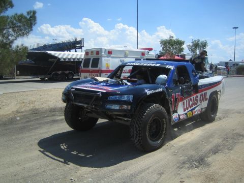 1990 Ford Bronco - blue number-17 trophy truck with Lucas Oil livery moves through a dirt pit area near an ambulance and race trailers.