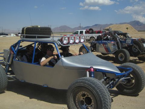 1990 Ford Bronco - silver tube-frame multi-seat off-road buggy carrying occupants and roof-mounted spares sits on a dirt lot with other race cars behind.