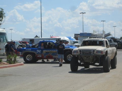 1990 Ford Bronco - tan prerunner pickup and several blue trophy trucks are staged in a paved pit lot with RVs and spectators under a cloudy sky.