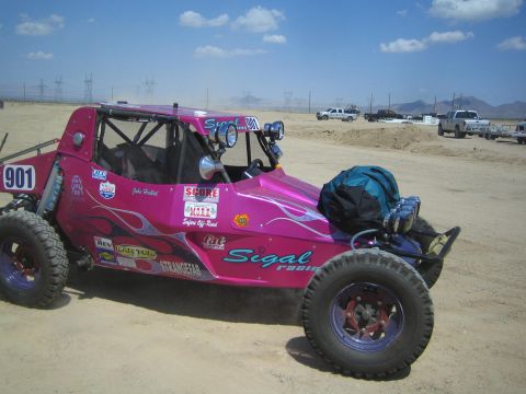 1990 Ford Bronco - pink number-901 tube-frame off-road buggy with Sigal Racing flame graphics and gear strapped on its rear sits on a desert dirt lot.