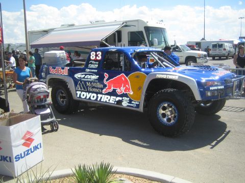 1990 Ford Bronco - blue number-83 Red Bull and Toyo Tires Chevrolet trophy truck sits in a paved pit with a motorhome, crowd and a Suzuki banner nearby.