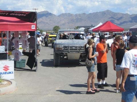 1990 Ford Bronco - mud-coated Ford race truck rolls through a desert off-road event midway lined with vendor canopies as spectators watch.