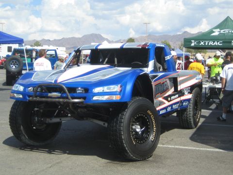 1990 Ford Bronco - blue-and-white off-road trophy truck numbered 20 sits on display near vendor tents with desert mountains behind.
