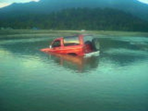 1985 Ford Bronco II - A red 1985 Ford Bronco II is partially submerged in water with a mountain and trees in the background.