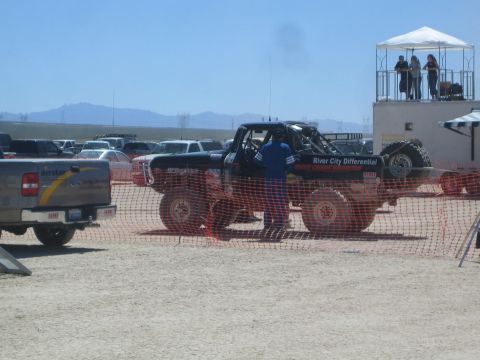 1990 Ford Bronco - black off-road race truck waits behind orange safety fencing in a dusty desert staging area beside a timing tower.