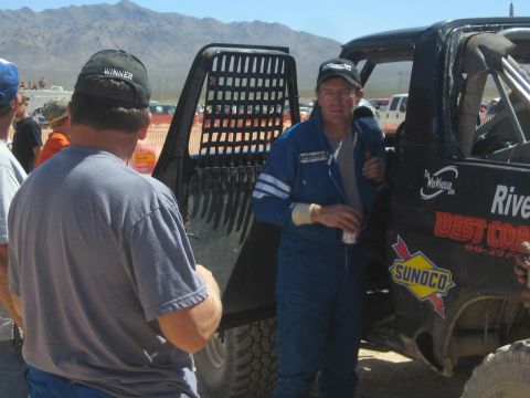 1990 Ford Bronco - man in a blue racing suit leans against a black off-road race truck talking with spectators in the desert.